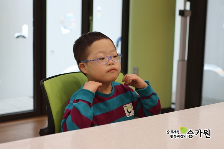 책상에 앉아 있는 김두영 장애아동의 사진. 보라색 안경을 쓰고  양손을 목 쪽에 올리며 앞을 응시하고있다 / 장애가족행복지킴이 승가원ci 삽입
