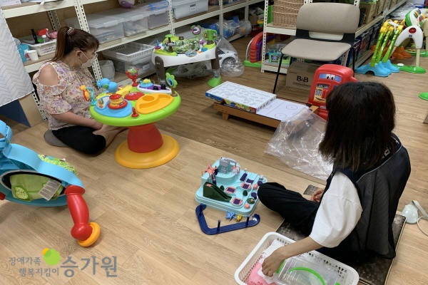 두명의 여성장애인근무자들이 자리에 앉아 아기 장난감을 살피며 근무 중에 있는 모습 주변에는 그외의 아기 장난감이 여러개 펼쳐져있/ 좌측 하단 장애가족행복지킴이 승가원 ci 로고 삽입