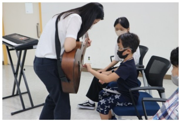 선생님이 기타를 들고 연주하고 있는데 장애아동이 줄을 만져보며 관심을 가지고 있다. 그 옆에는 자원봉사자가 앉아있다.
