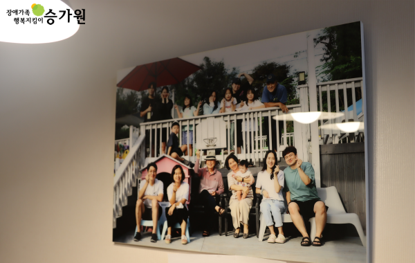 왼쪽 상단에 장애가족 행복지킴이 승가원ci / 열여섯명의 대가족이 윗층 아랫층에 나뉘어 앉거나 서서 찍은 사진이 하얀 벽에 걸려있음.
