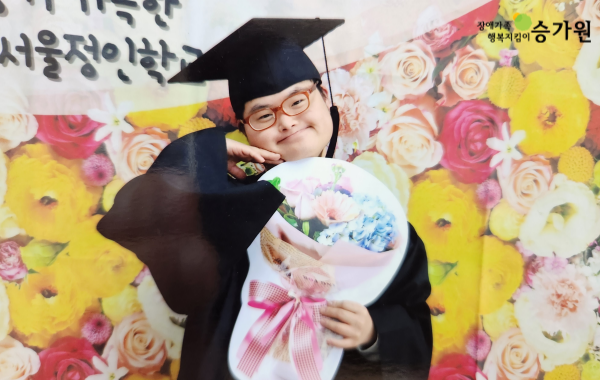 가득한 서울정인학교라고 쓰인 꽃이 가득한 현수막 앞으로 졸업모자를 쓰고 분홍색 꽃다발을 들고있는 남성 / 오른쪽 상단 장애가족 행복지킴이 ci