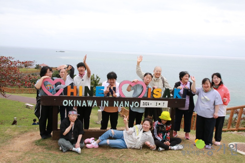 12명의 장애가족이 OKINAWA IN NANJO라고 적힌 팻말에 둘러 사진을 찍고 있는 모습/ 주위는 큰 들판으로 들판 뒤 바다가 보인다./ 우측 하단 승가원CI삽입