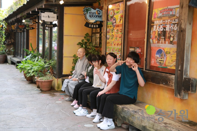 가게 앞 나무 의자에 4명의 장애가족과 스님이 앉아 사진을 찍고 있다. 우측하단 승가원CI삽입