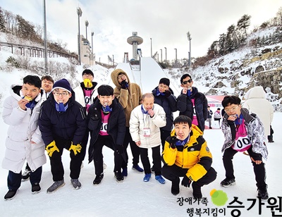 눈이 덮인 경기장에서 11명의 성북장애인복지관 선수들이 메달을 입에 물고 다같이 사진을 찍고 있는 모습. 오른쪽 아래 장애가족 행복지킴이 승가원CI